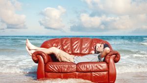 Ein Mann liegt entspannt auf einem roten Sofa am Strand.