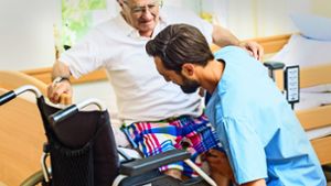 Ein Pfleger hilft einem Senior vom Rollstuhl ins Bett. Foto: Kzenon/stock.adobe.com